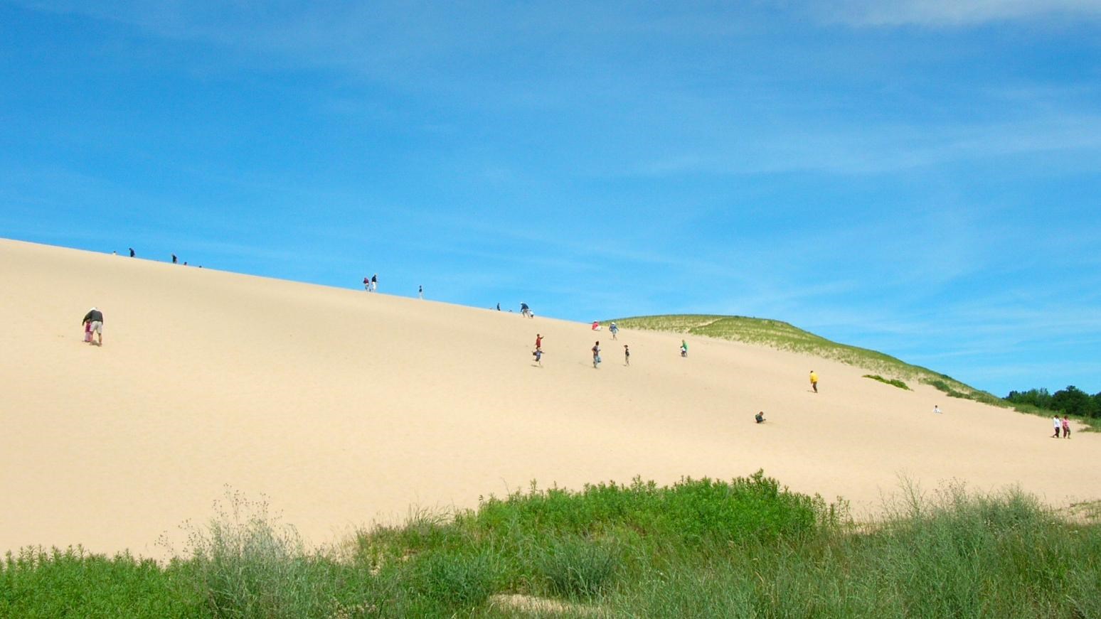 A large sand hill against a azure sky with green dune grass in the foreground