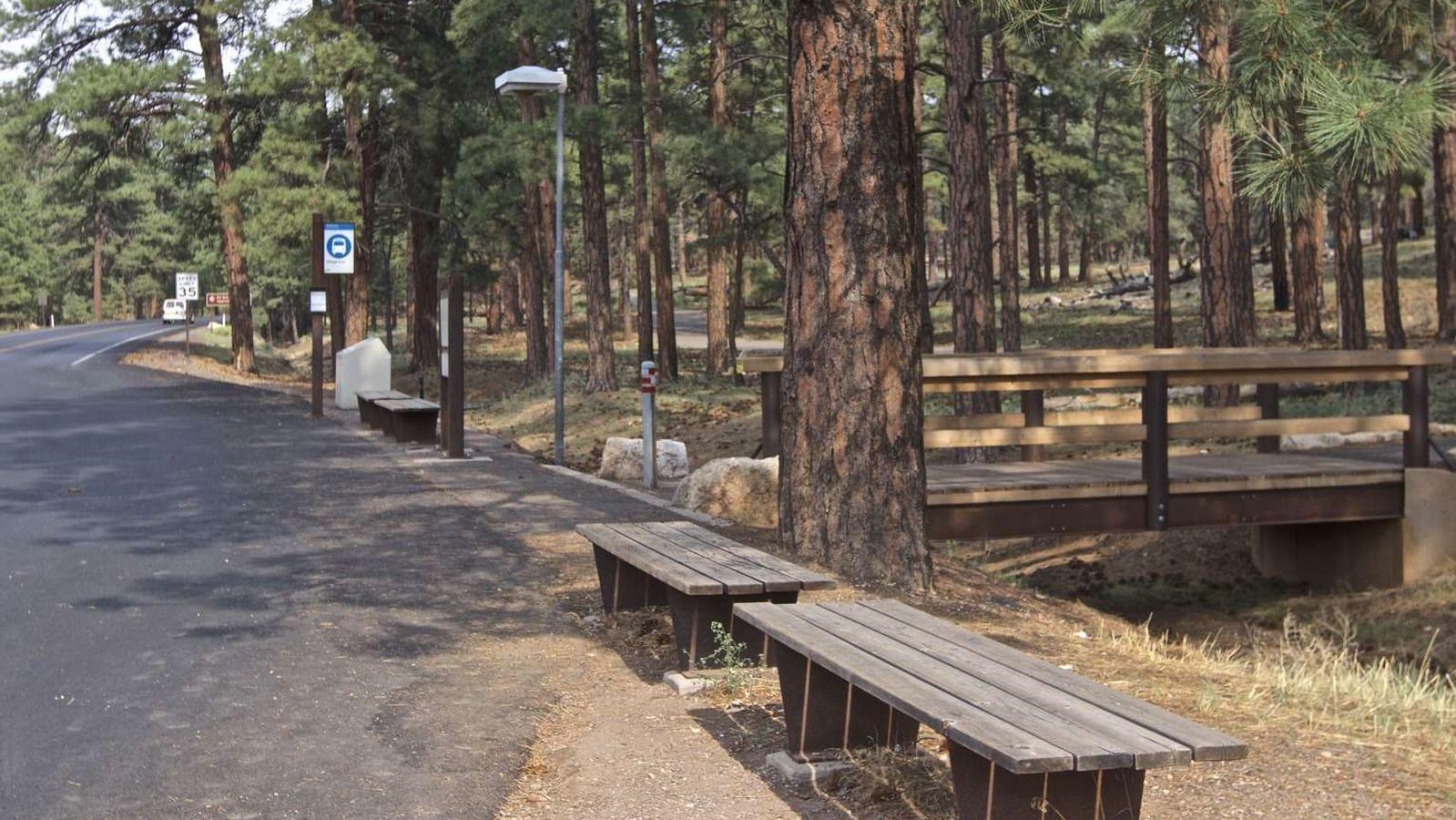 A wooden benches next to a shuttle bus stop. A wooden footbridge in the background.