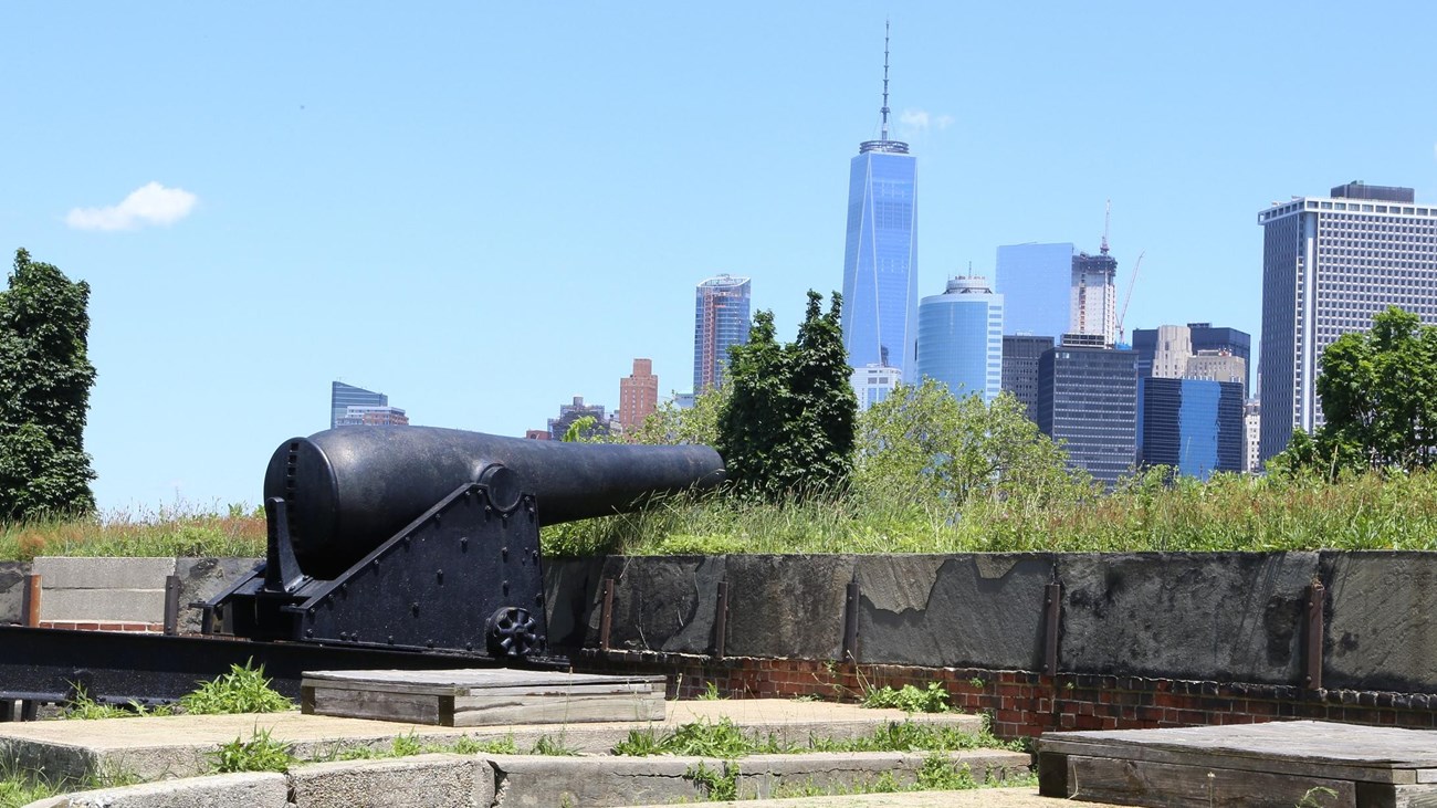 large, black, iron cannon pointing over forts wall with NYC skyline in backround