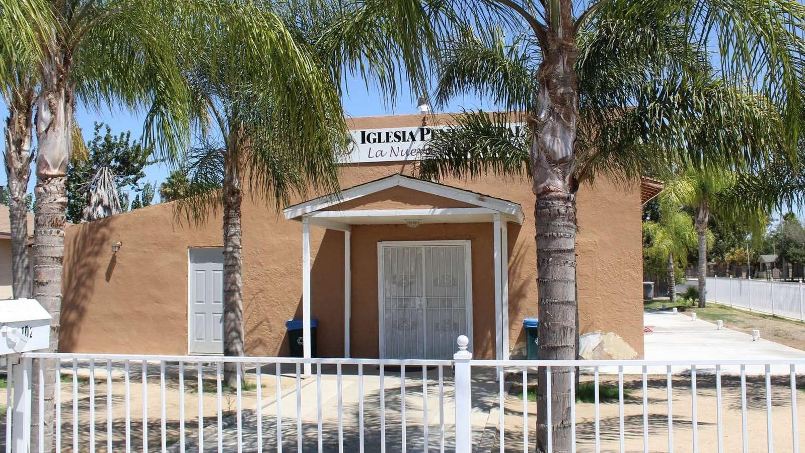 An orange adobe building with white trim is behind trees and a white fence. A sign is obscured.