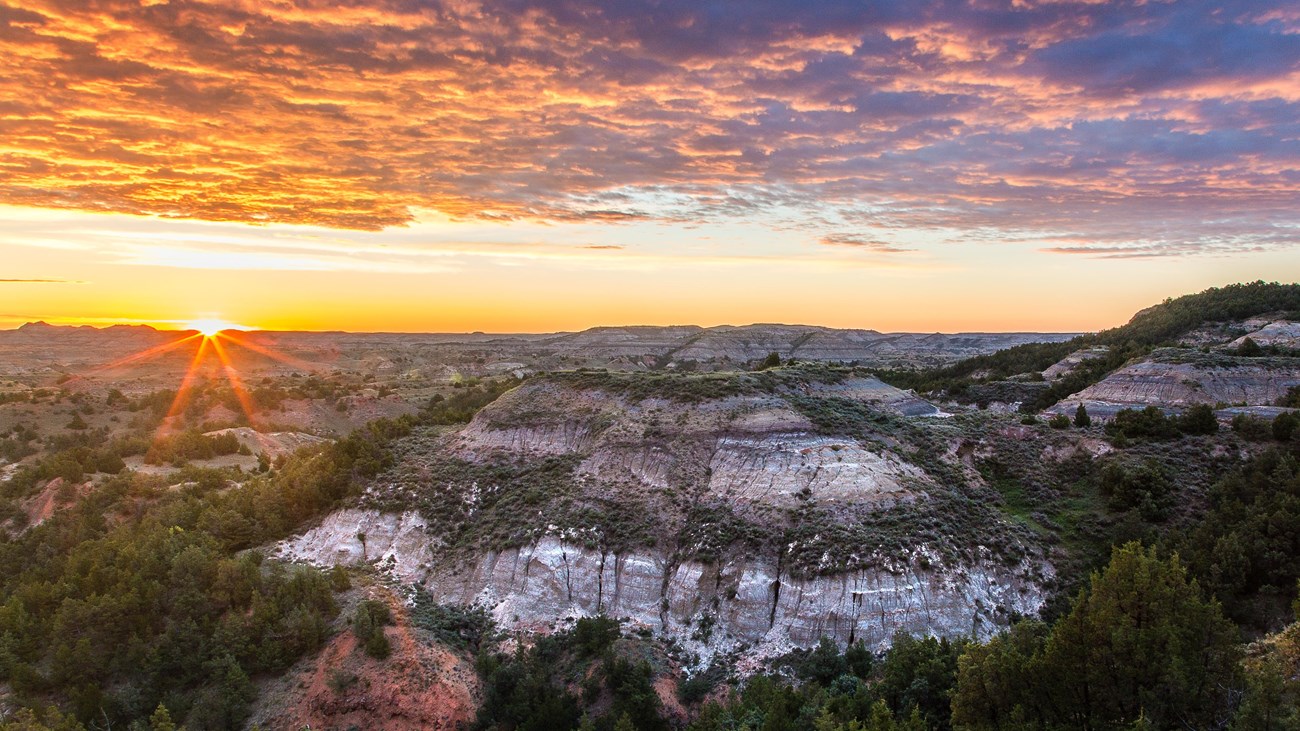 The sun is just visible over a badlands scene. Juniper grow on the buttes, and there are low clouds.