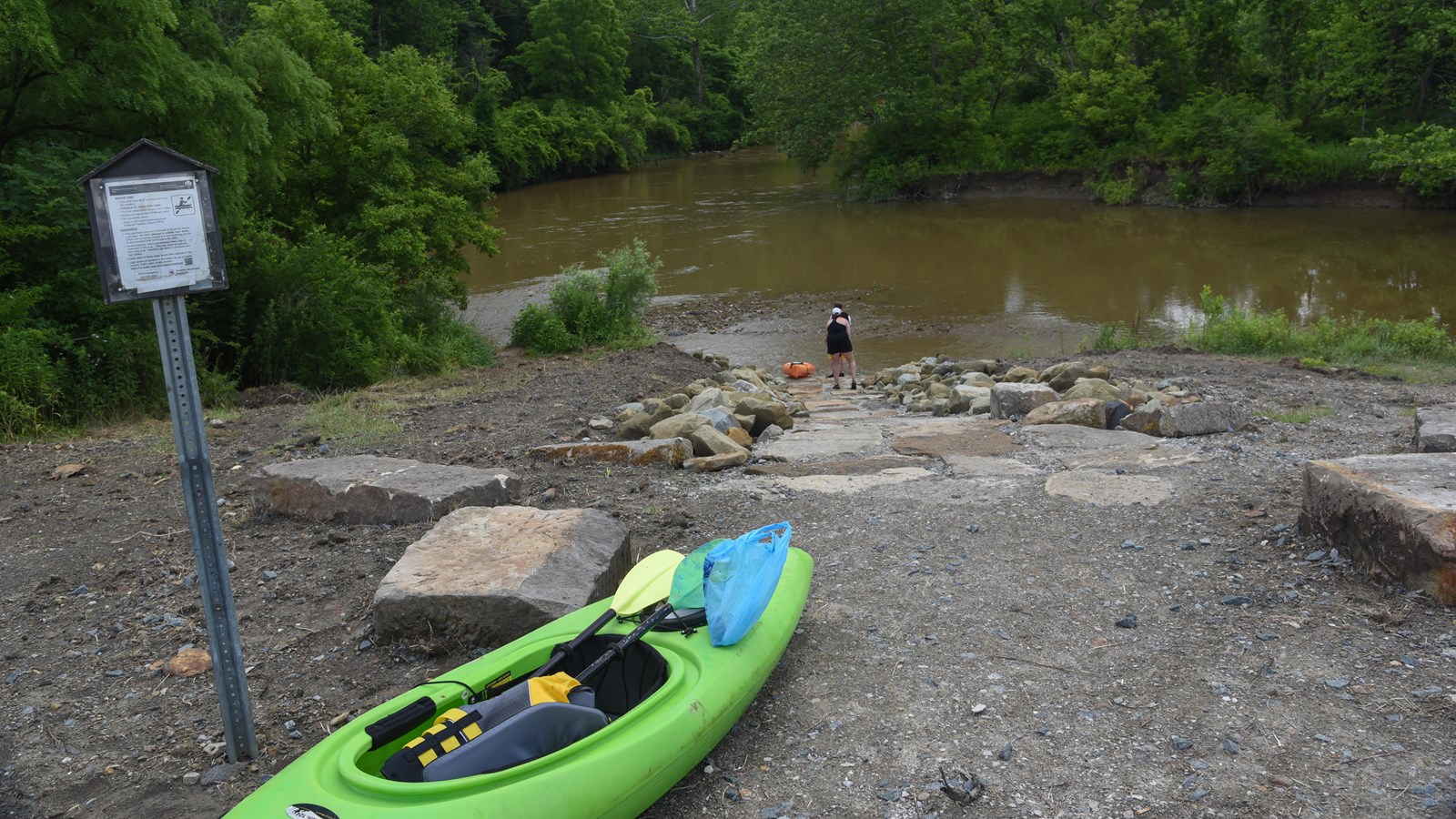A green kayak rests by a trail sign overlooking the junction of two waterways. Two people pull an or