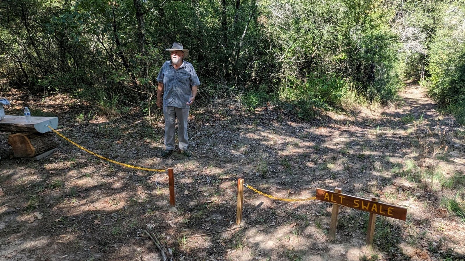 A man stands next to a dirt trail with a sign indicating a trail swale.