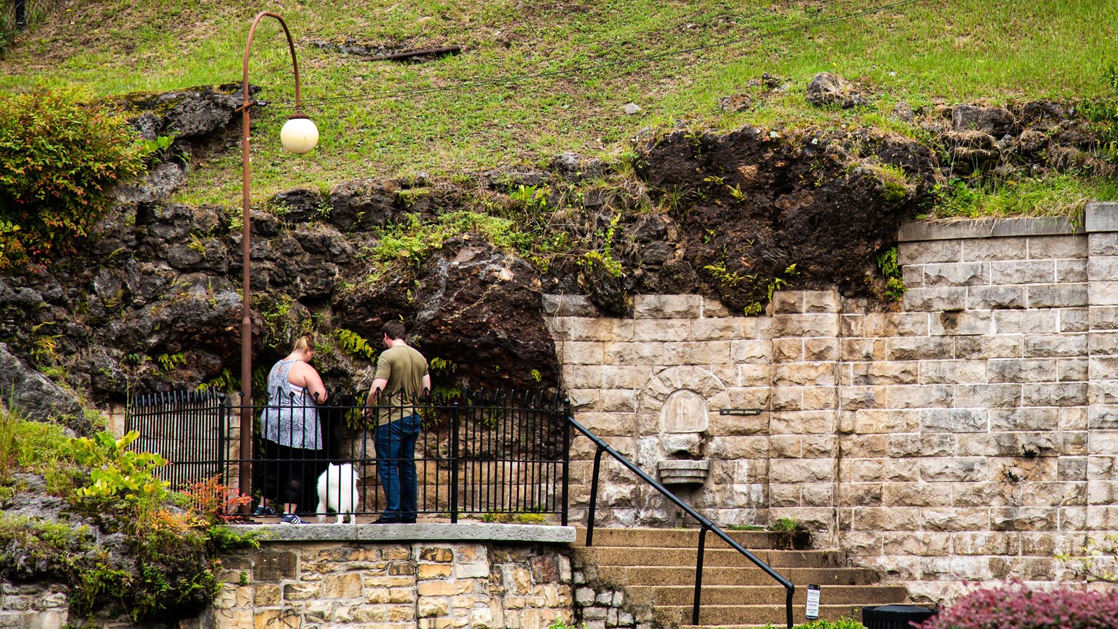 A couple and their dog stand on the top of a few stairs peering into a spring in a tan brick wall.