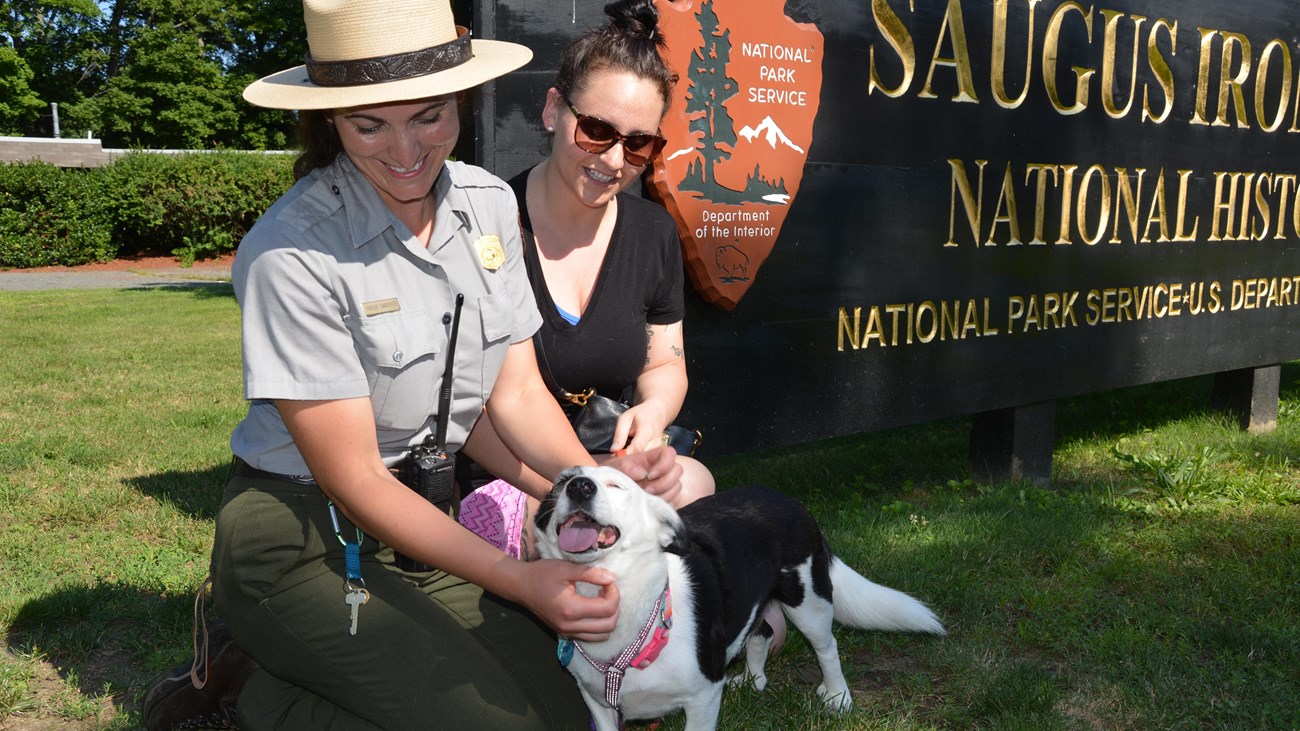 Two people sit on the grass petting a black and white dog in front of the Saugus Iron Works sign
