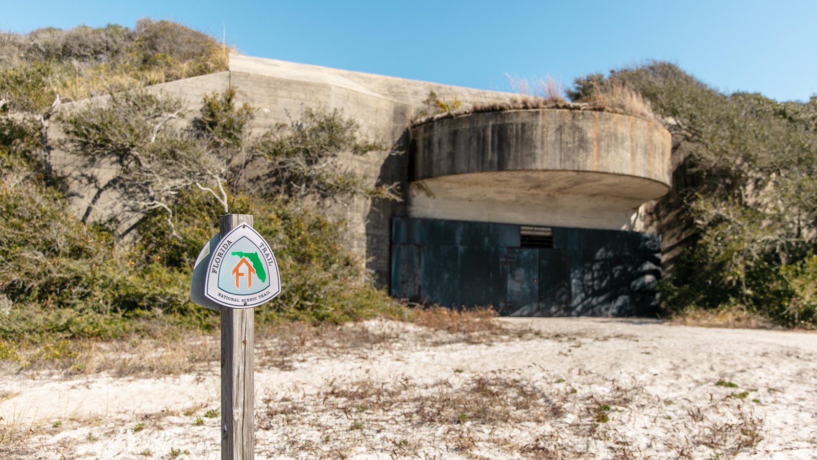 A sign for the Florida National Scenic Trail stands in front of a concrete battery with black doors.