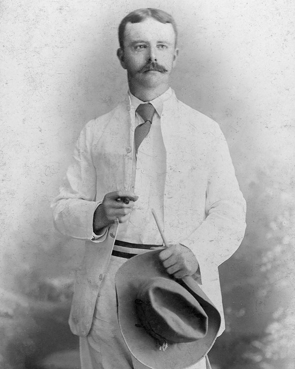 A man wearing a white suit, holding a hat and a cigar.