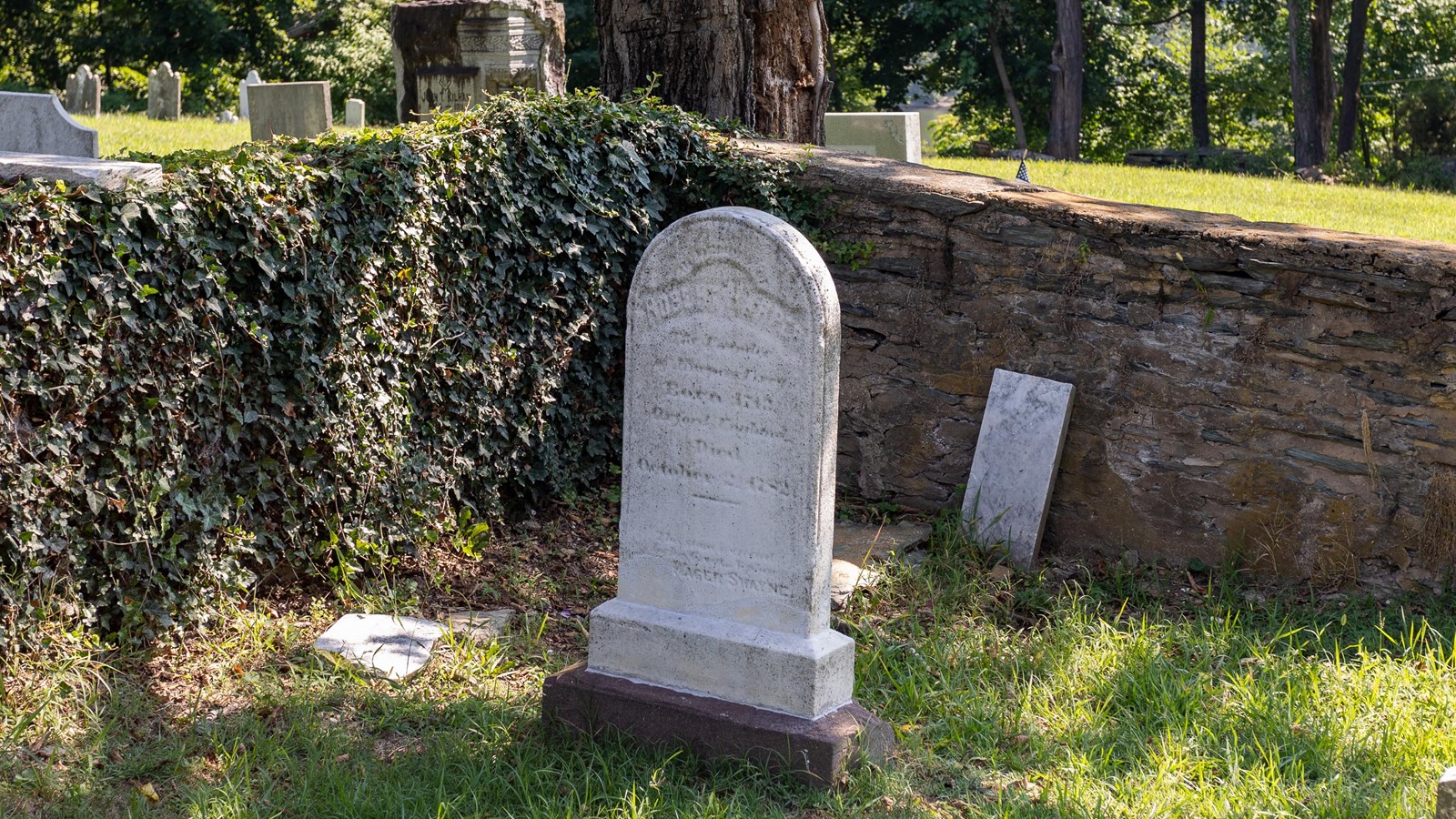 A headstone stands at the corner of an enclosure surrounded by a stone wall, underneath a tree.