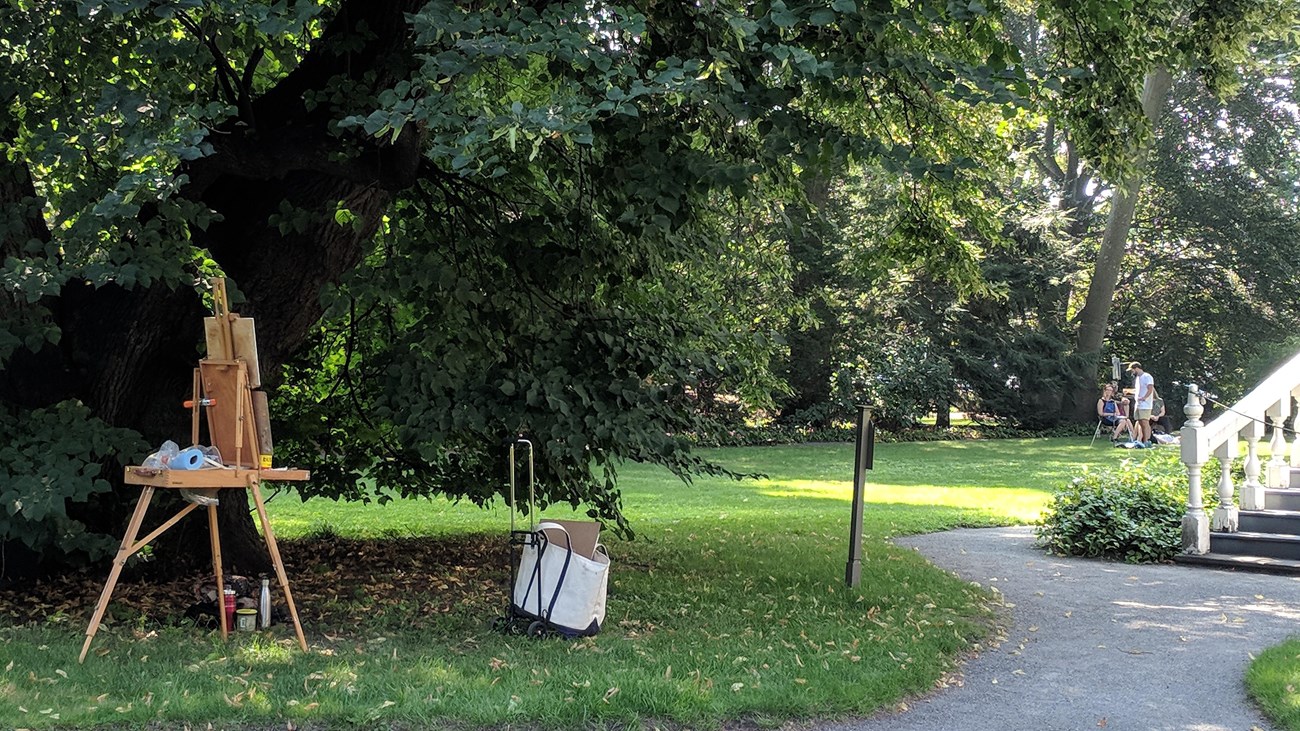 artist\'s easel set up under large tree, with group of people around second easel on lawn behind