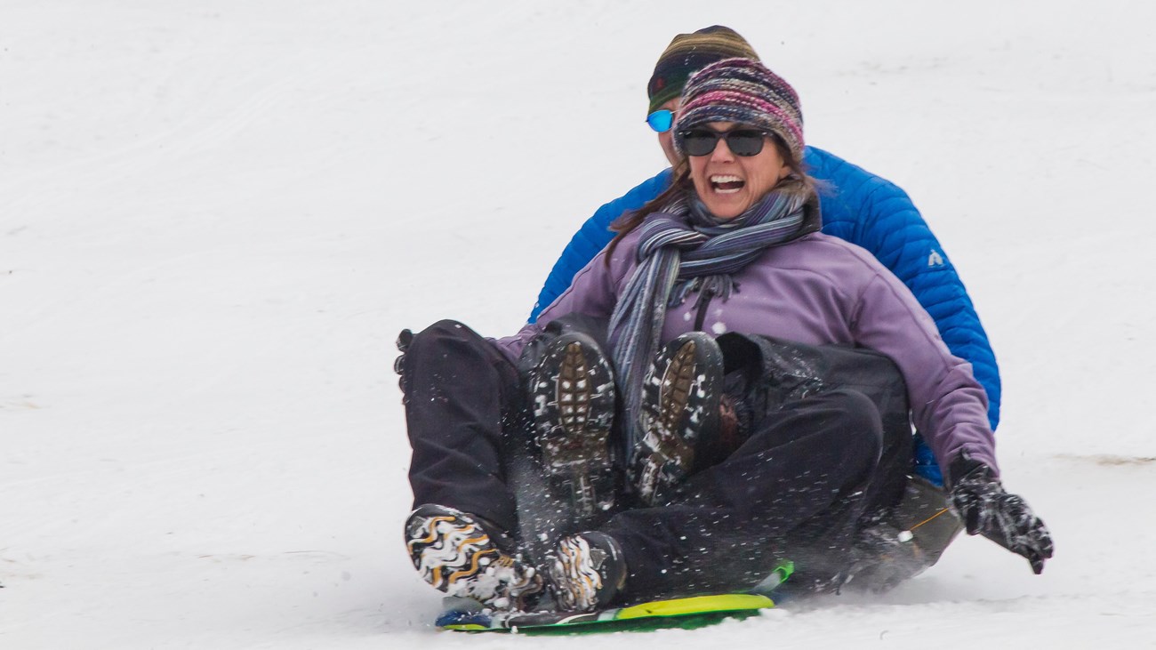 Two visitors are sitting on a sled and sledding down a snowy hill