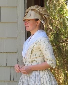 Woman in 18th century light colored gown and bonnet stands in a doorway
