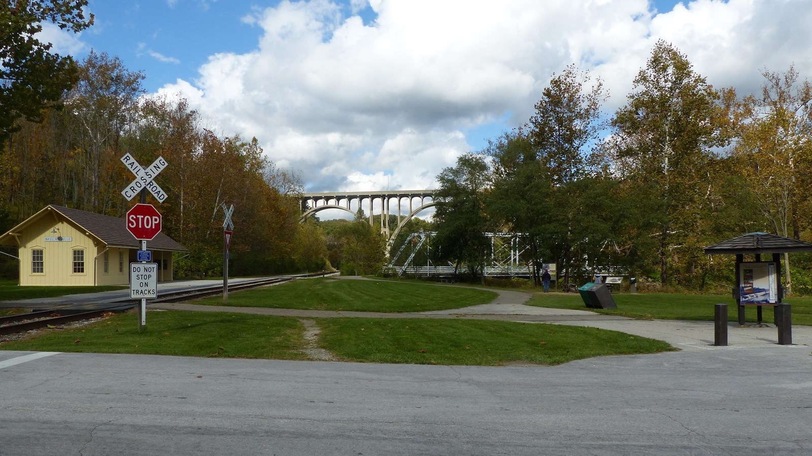 Yellow train station and tracks to the left of a distant arched bridge. Three-sided kiosk at right.