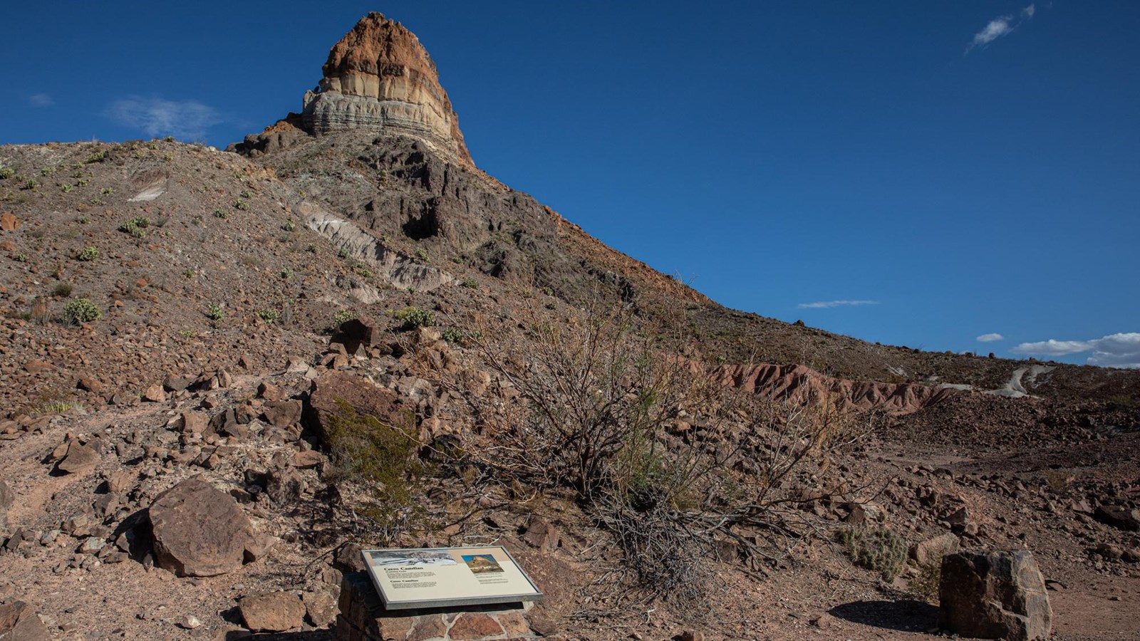 A metal sign atop a rock pedestal is located at the base of a volcanic mountain.
