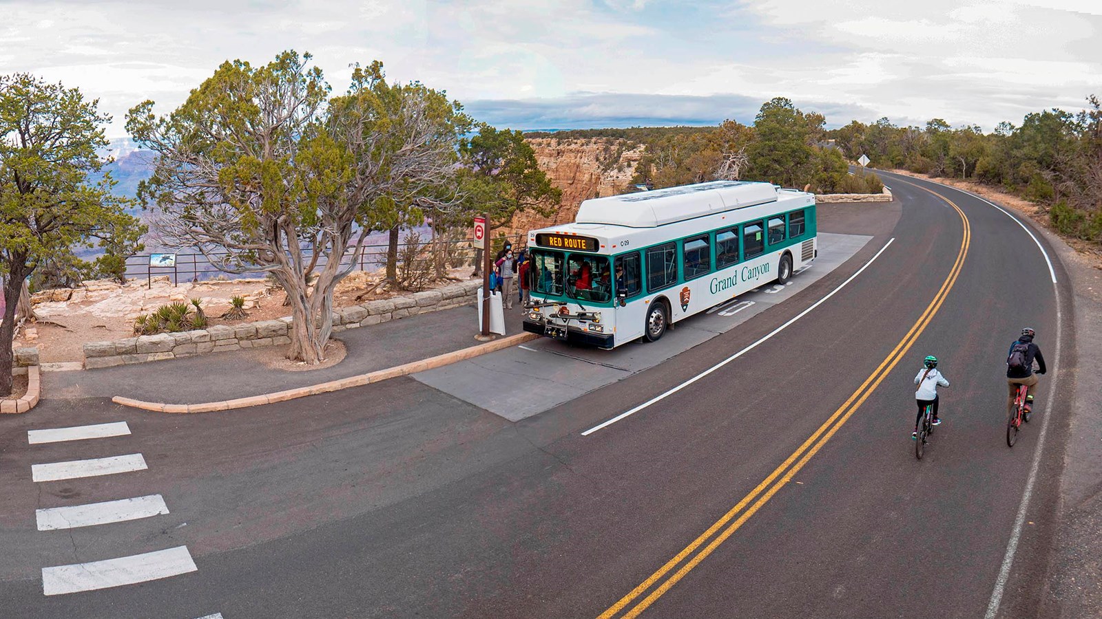 a white and green bus is parked at a stop along the canyon rim. Two people ride by on bicycles.