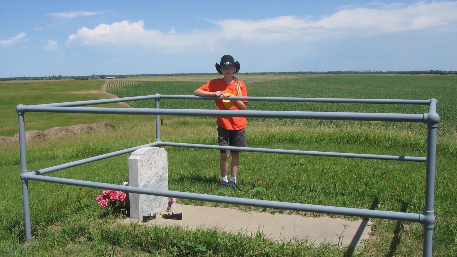 A person stands behind a grave enclosed by a rail fence in a grassy landscape.