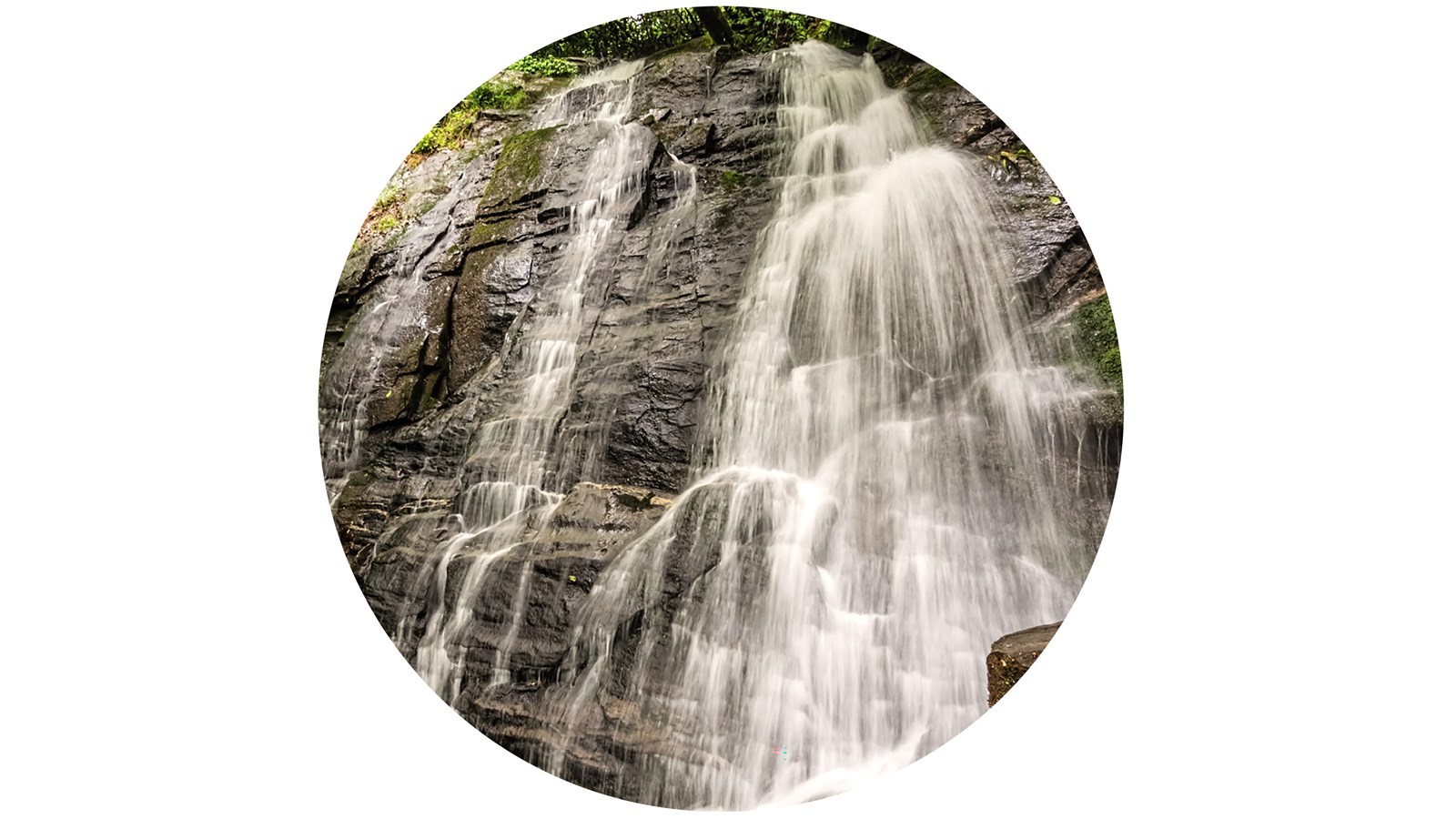 A fanning waterfall flowing over rocks framed by shrubs and trees.