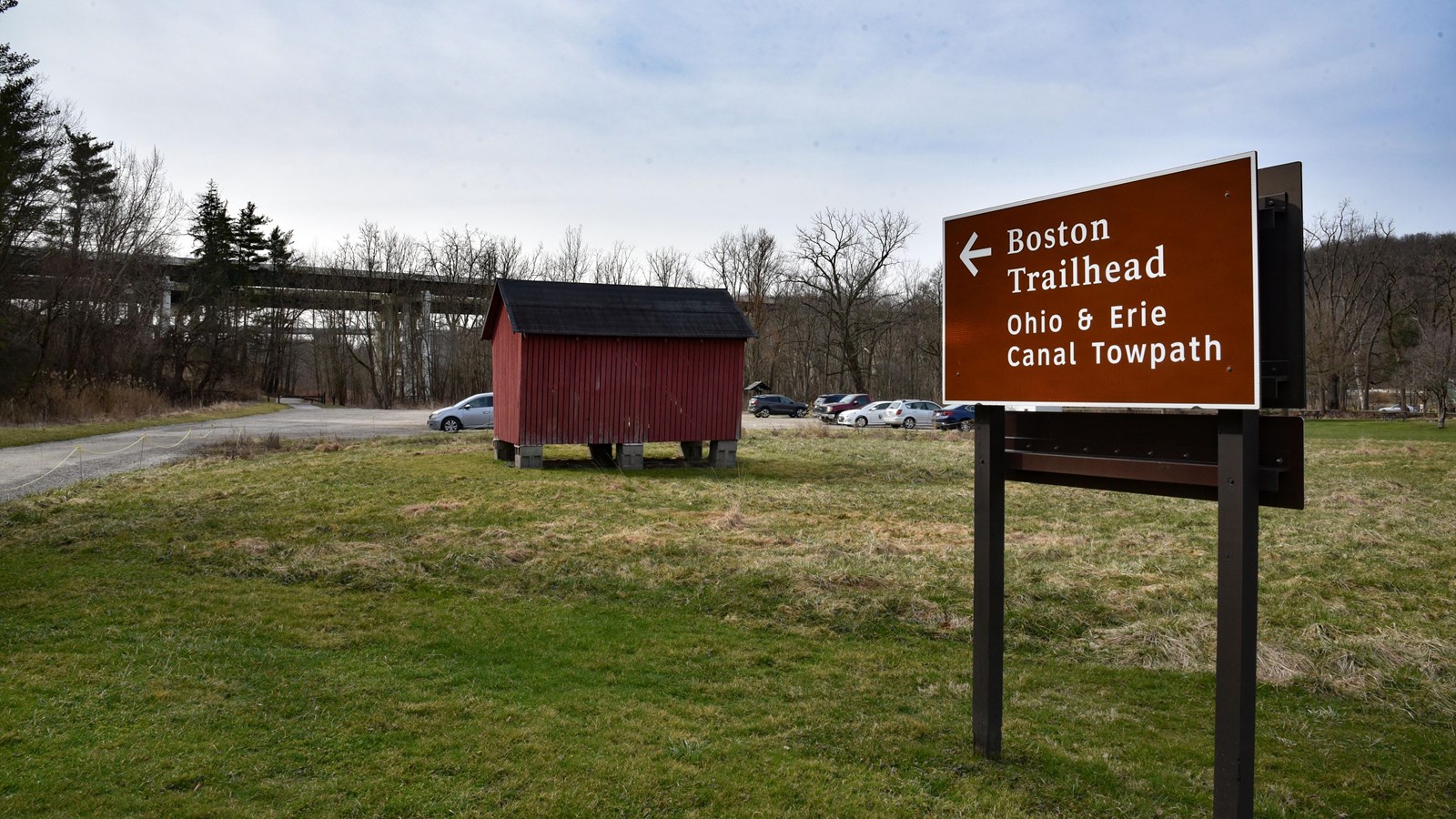 Road sign points to unpaved parking lot with a red corn crib, center, and distant highway bridges.