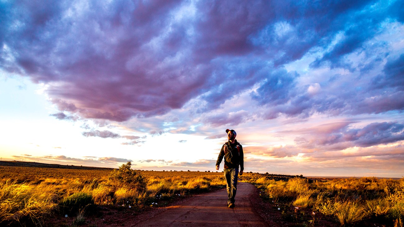 Man walking a trail under a sunset sky with clouds