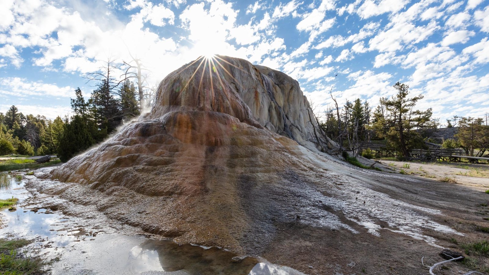 Steaming water flows down a terraced rock formation covered in orange bacterial mats.