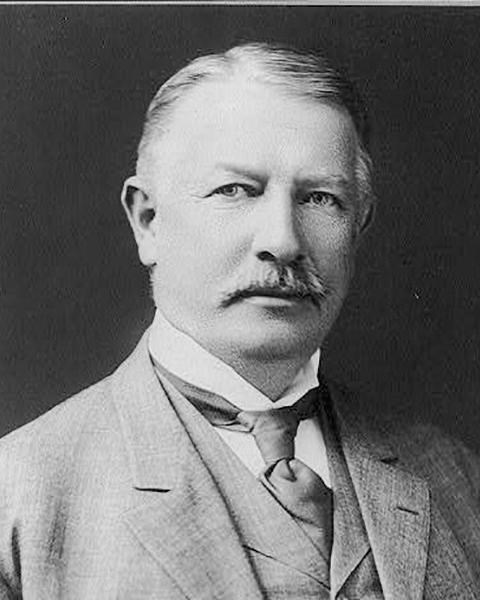 black and white photo of a man with a mustache. He's wearing a coat with a vest and high collar shir