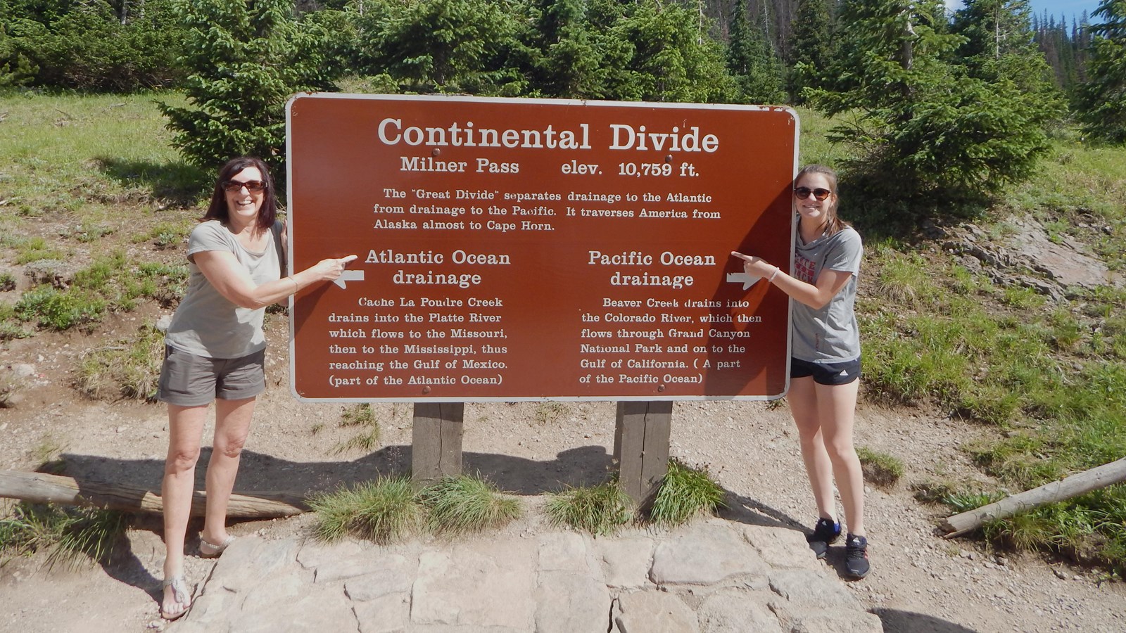 Two people stand next to a brown sign that reads: Continental Divide, Milner Pass, elevation 10,759 