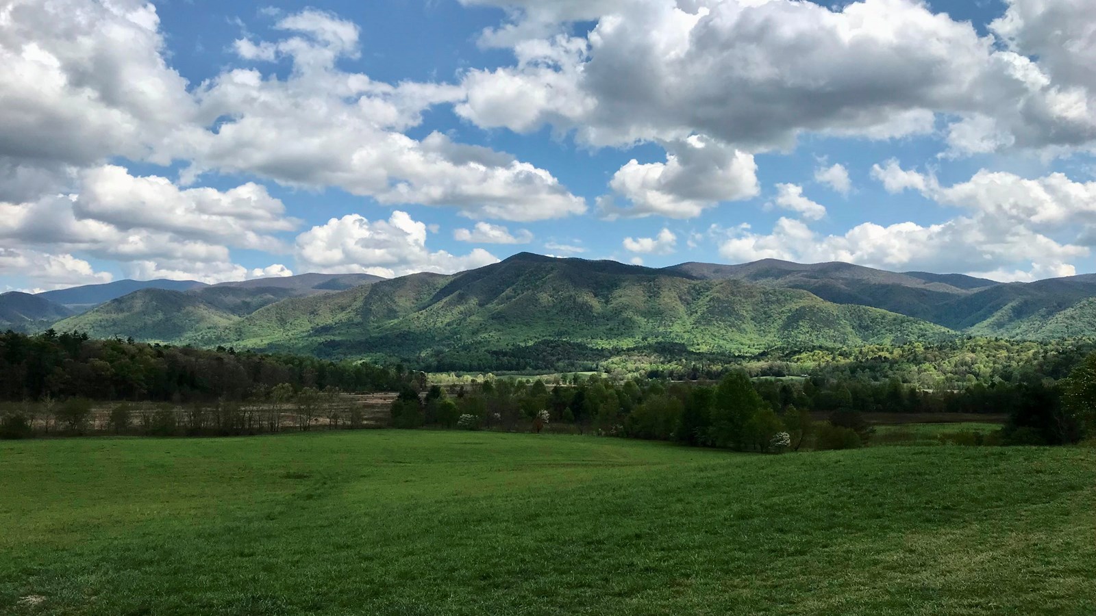 A grassy valley surrounded by distant mountains with blue sky and clouds above.