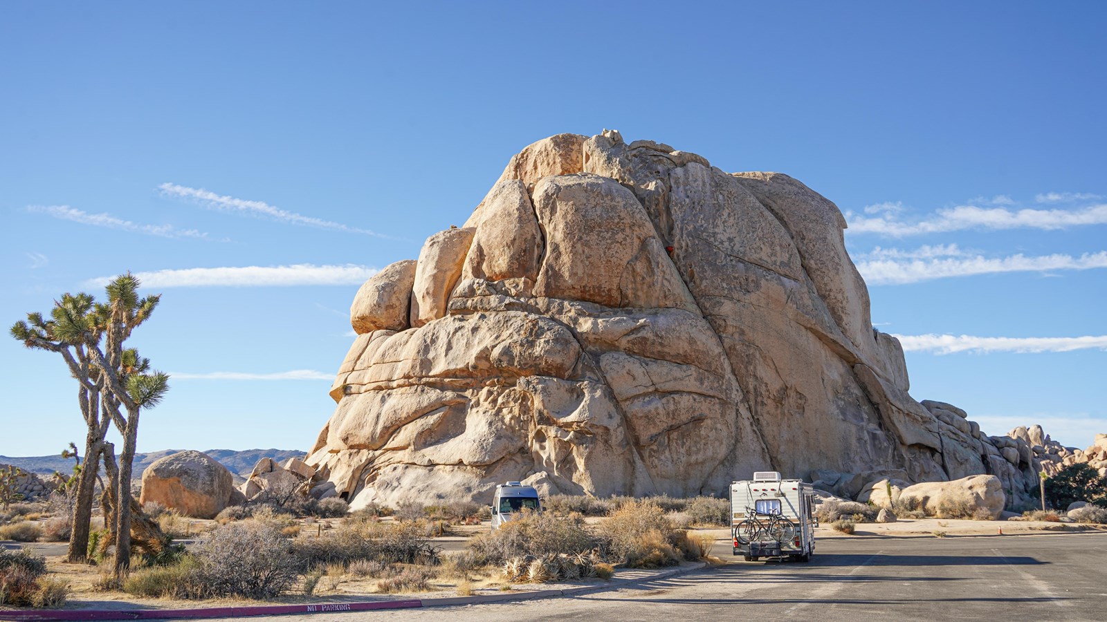 A large boulder sits behind a parking area with two vehicles. There are Joshua trees to the left.