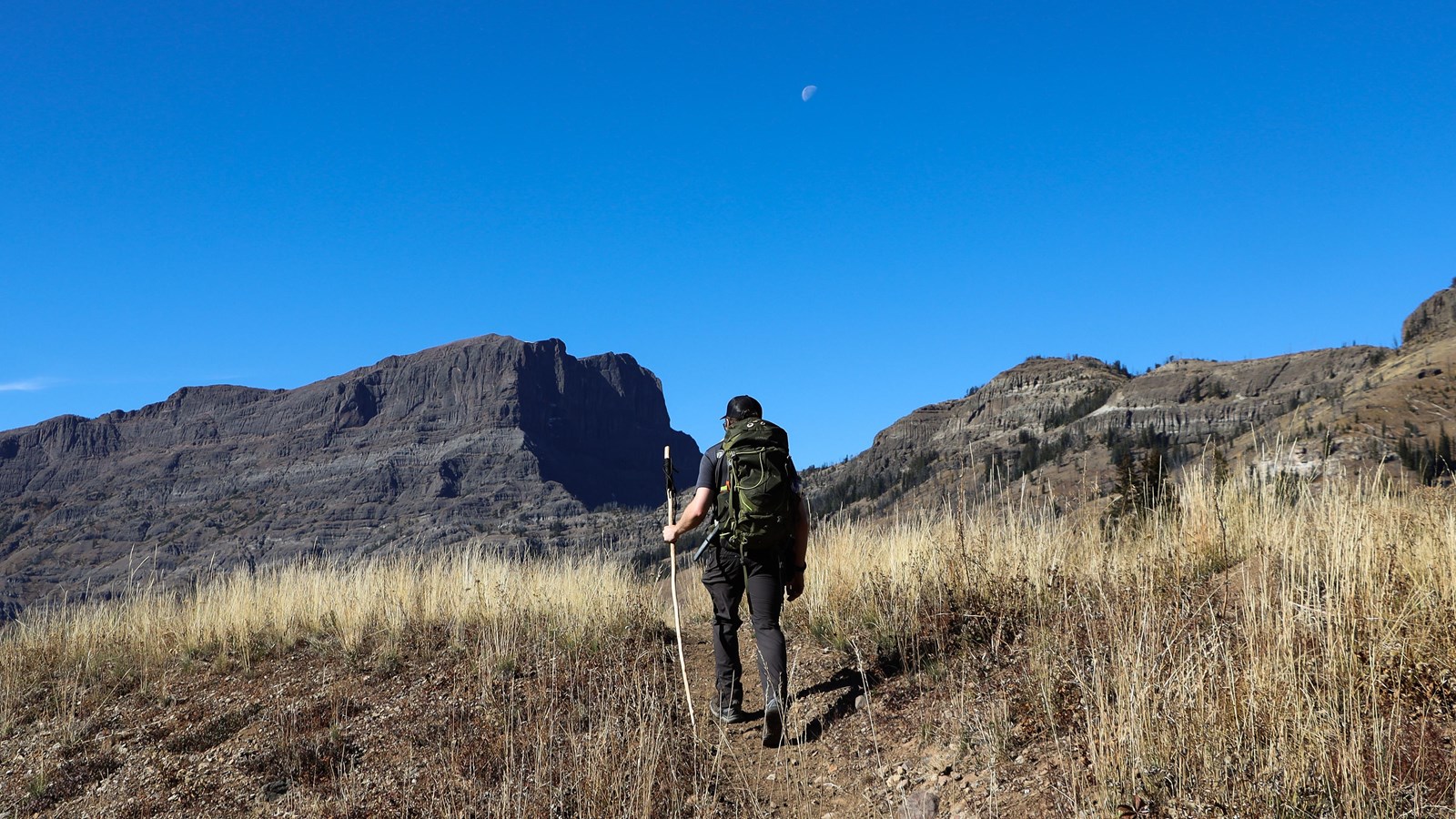 A person hikes along a trail through dry vegetation beneath rocky cliffs.