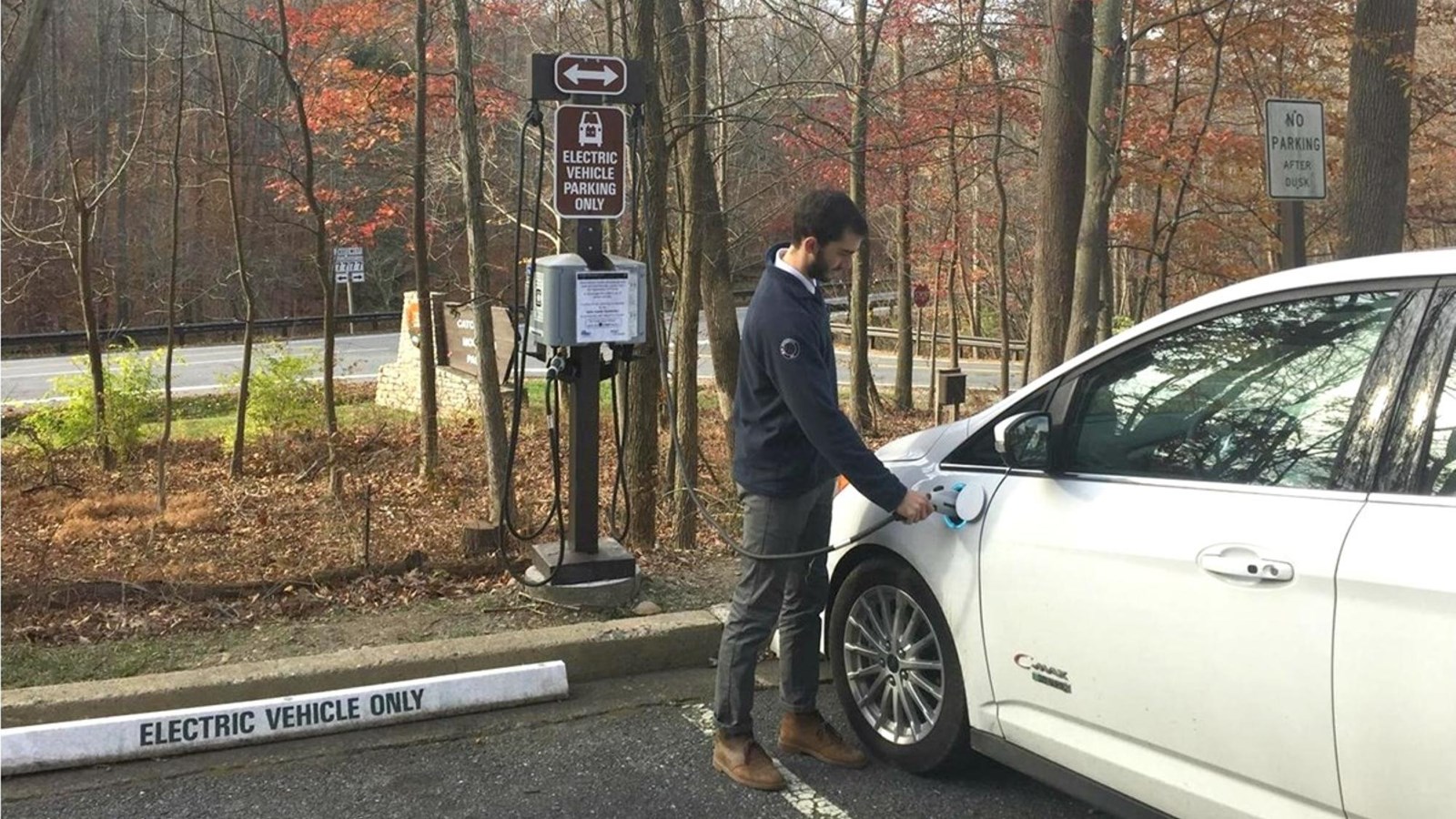 A man plugging in a power cable from an electric vehicle power station to a white car.
