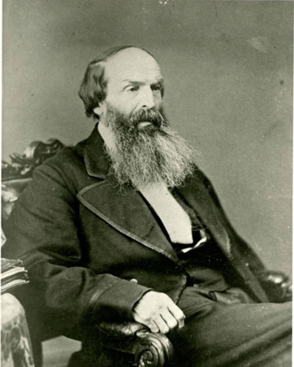 A black and white portrait of a man with short hair and long beard seated in a chair wearing a suit