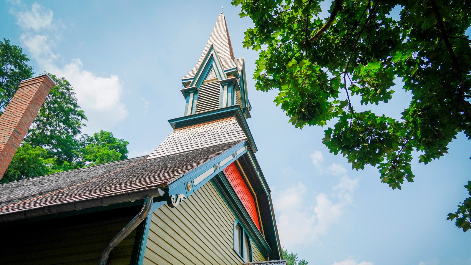 A side view of a green and orange church building with a tall steeple and a tree in front.