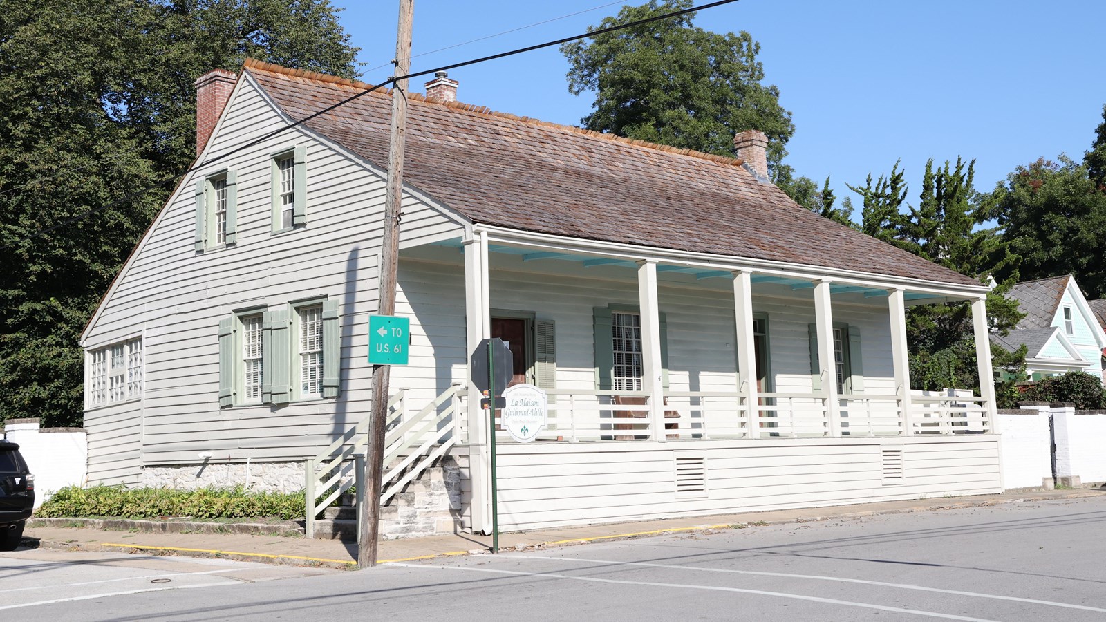 Angled view of a grey house with green shutters with front and back porch and brown shingled roof.