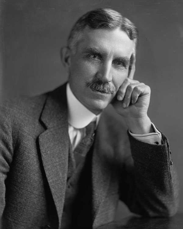 Portrait of a man with a mustache. His elbow is on a table and his face rests on his hand