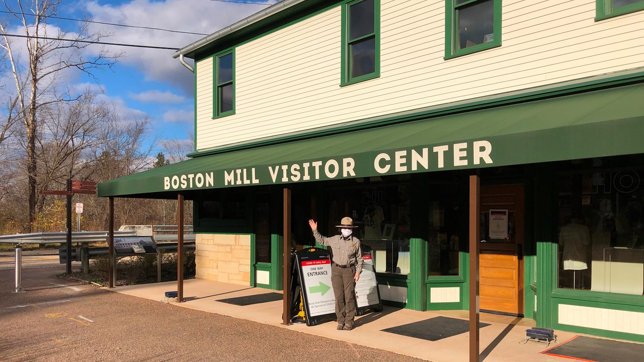 Female ranger wearing a mask and uniform waves hello on porch of a cream building with green awning.