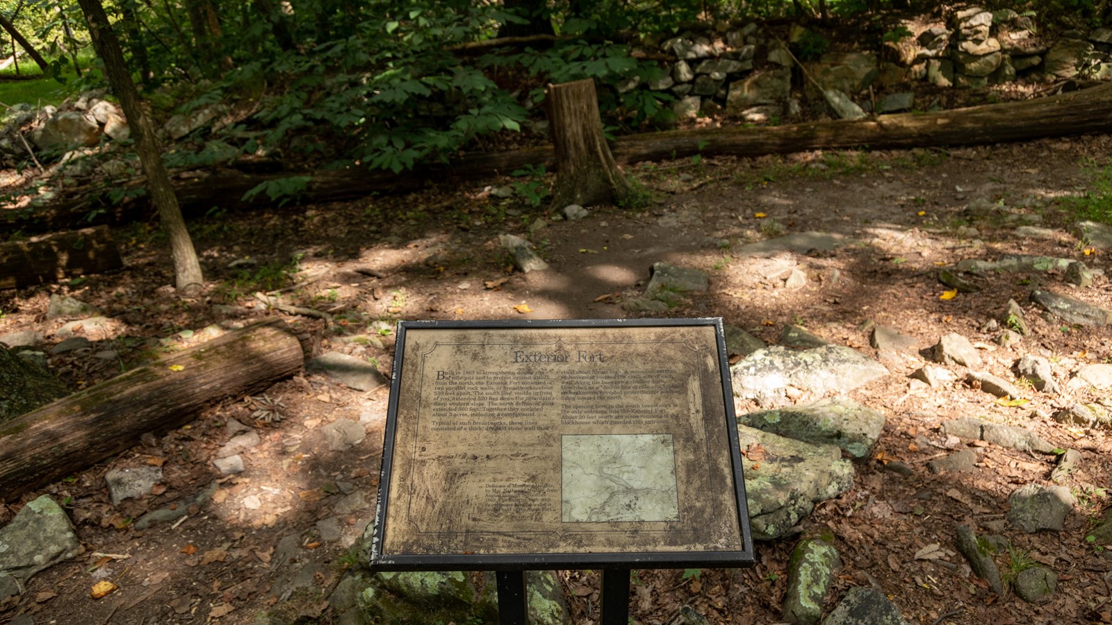 A sign sits in front of an overgrown area surrounded by a low rock wall.