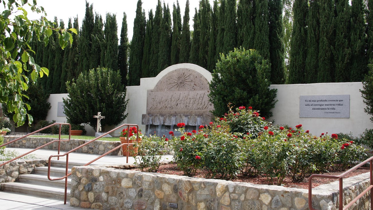 A bed of roses in front of a large wall fountain and grassy area around a grave marker