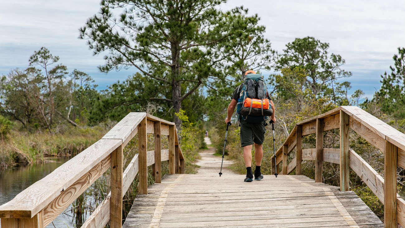 A backpacker crosses over a bridge with poles in hand.