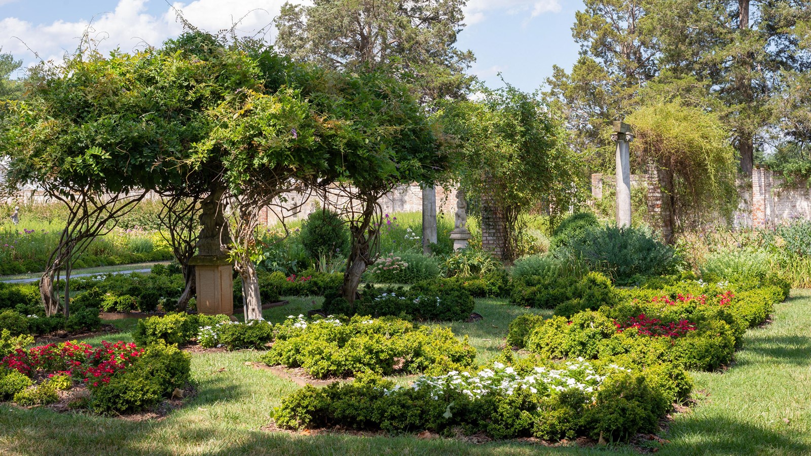 View of a walled, lush garden, with flower beds, a cherub statue, and pillar features..