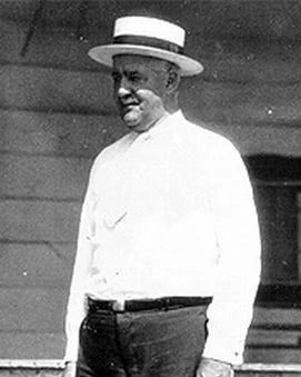 A black a white photo of a man standing. He is wearing a white shirt, black pants and a straw hat.