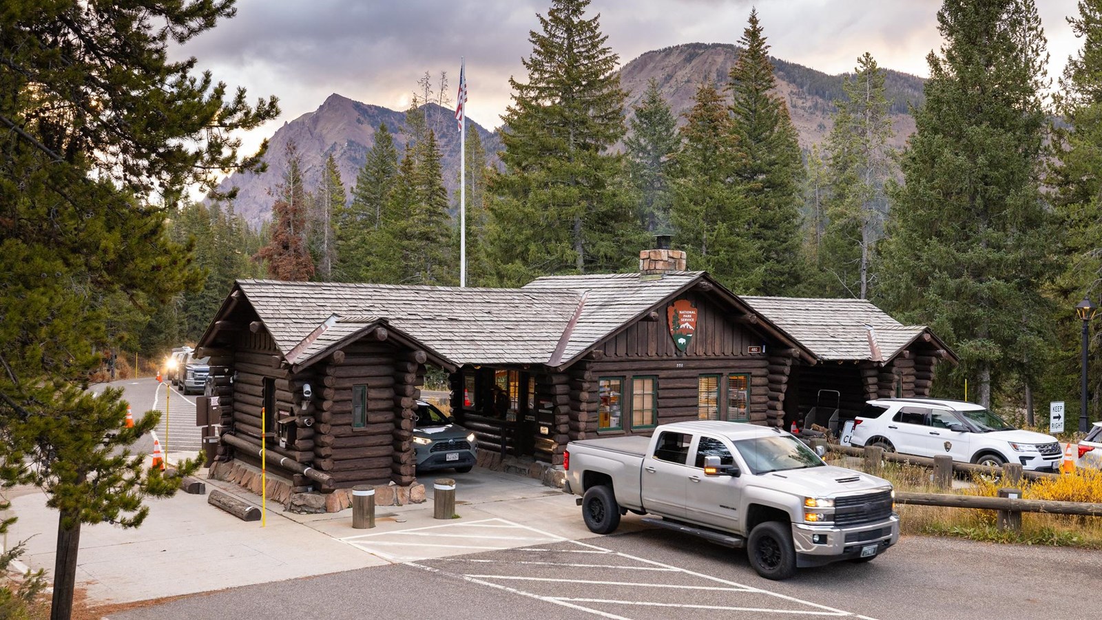 Vehicles drive through a historic entrance station made of logs in a forest with mountains.