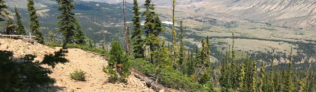 View of a valley covered in sagebrush-steppe from a tan, rocky trail near the top of a mountain.