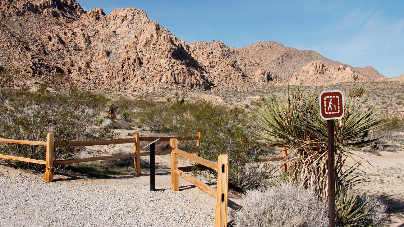 A dirt trail starting between wooden fences with rocky mountains in the distance.