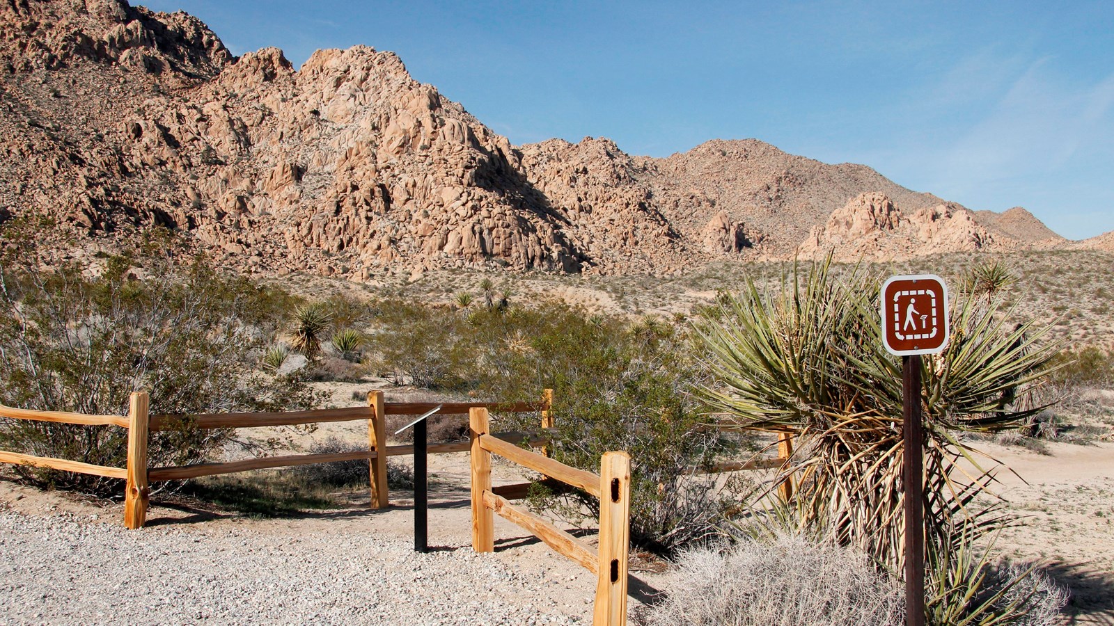 A dirt trail heading between two wooden split rail fences with rocky mountains in the background.