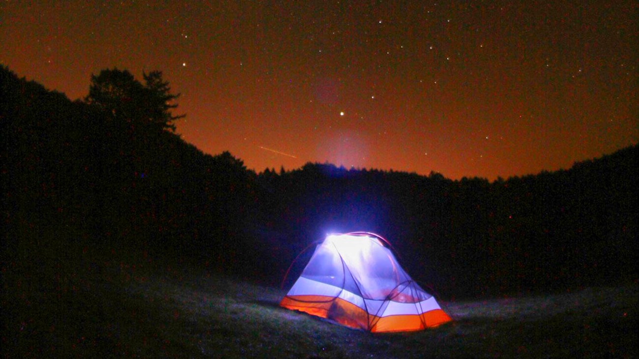 An illuminated two-person tent under a starry sky with orange light pollution on the horizon. 