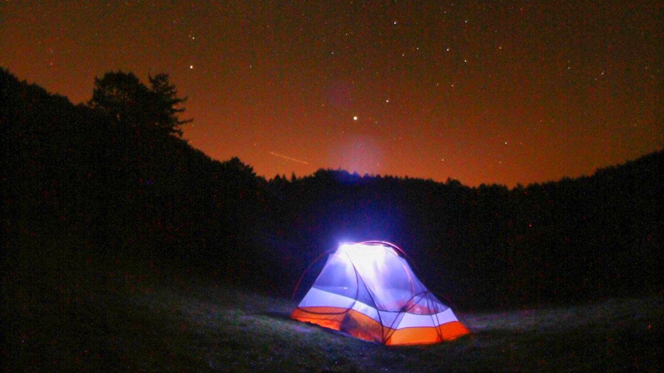 An illuminated two-person tent under a starry sky with orange light pollution on the horizon.