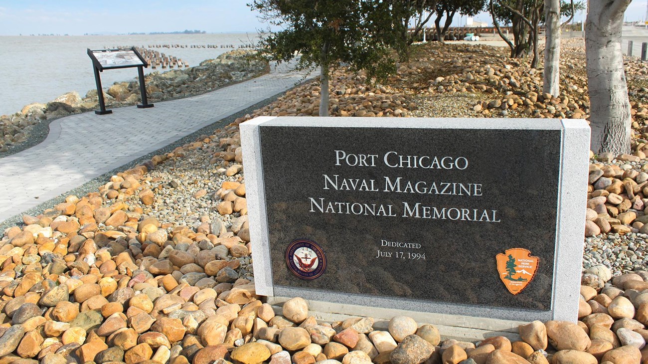 A park entrance sign in foreground. A pathway meanders along the waterfront. 