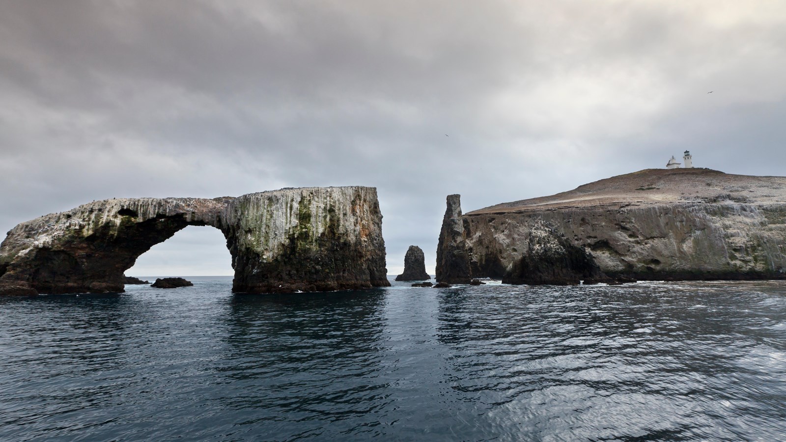 Steep dark volcanic rock has been eroded into a 40-foot-high arch that is surrounded by water. Behin