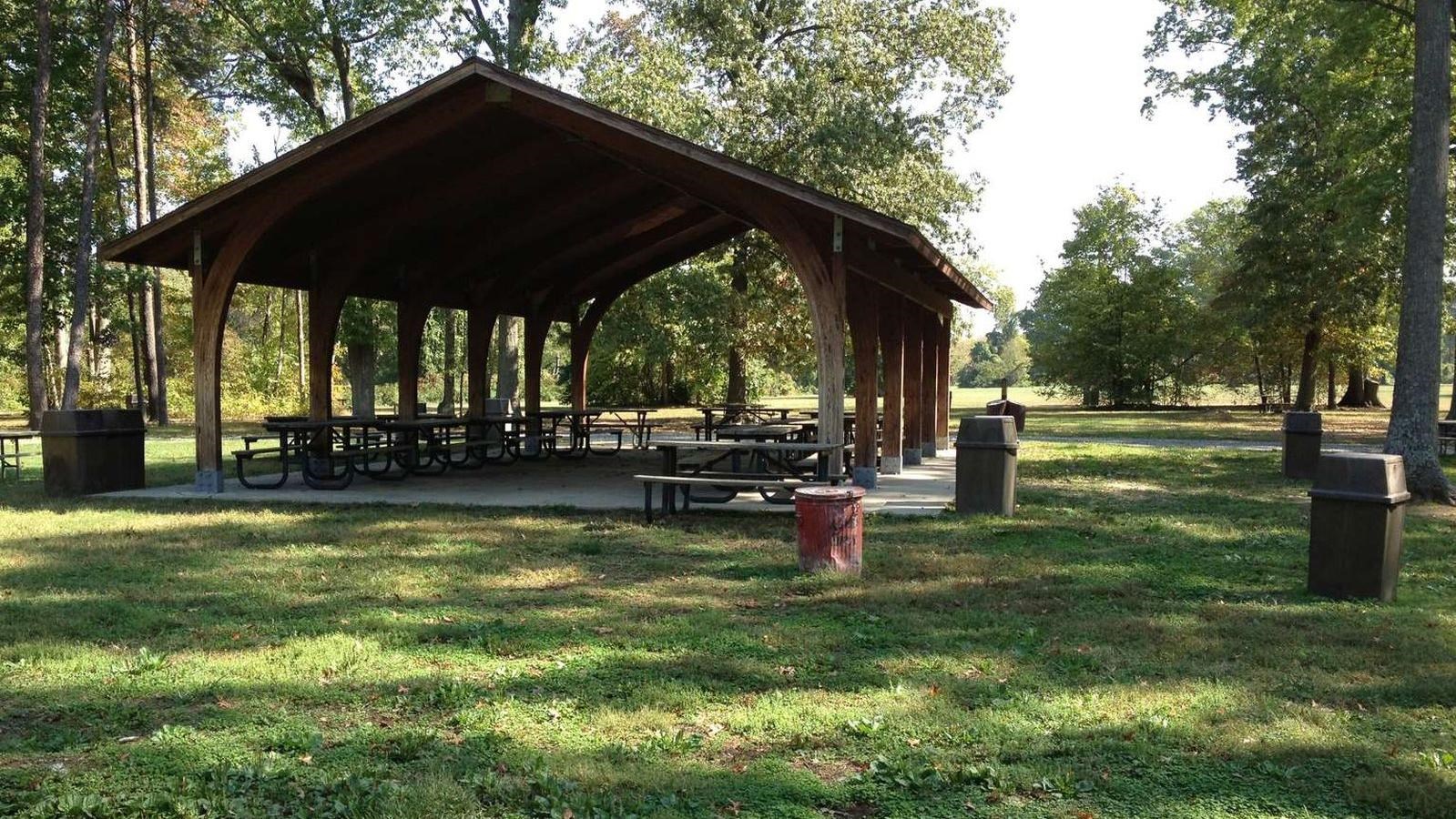 A wooden picnic pavilion structure covers picnic tables in a grassy field with trees in the summer
