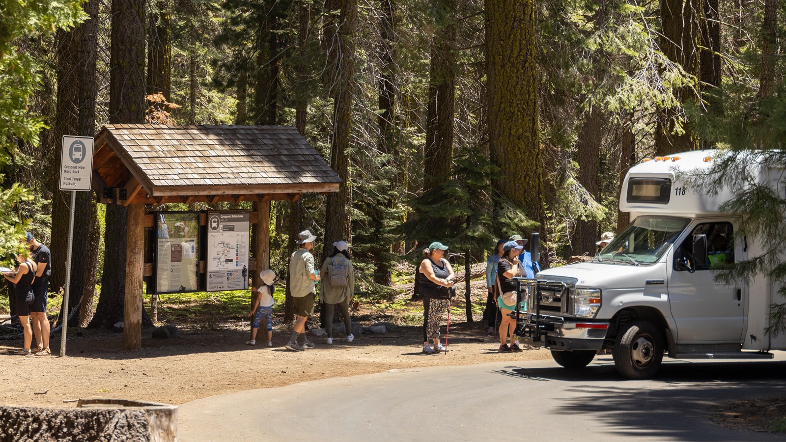 Park visitors stand next to a white shuttle van next to a wooden kiosk labeled