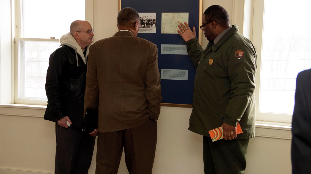 A ranger in a green jacket points to a photo for two guests standing beside him